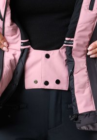 Schwarze und rosa Skijacke mit einem Innenfutter, das einen rosa Innenschlapf mit zwei schwarzen Druckknöpfen, schwarzem Futter und verstellbarem Kabelzug enthält.