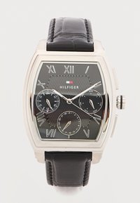 Τετράγωνο ασημένιο ρολόι με μαύρο καντράν, ρωμαϊκούς αριθμούς, τρεις υποκαντράν, μαύρο δερμάτινο λουράκι και λογότυπο Hilfiger στο κέντρο.