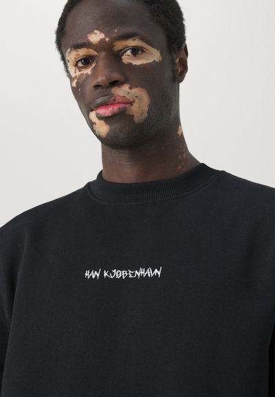 Black sweatshirt with ribbed neckline. Features white embroidered text "HAN KJØBENHAVN" on the chest. Smooth texture, casual fit.