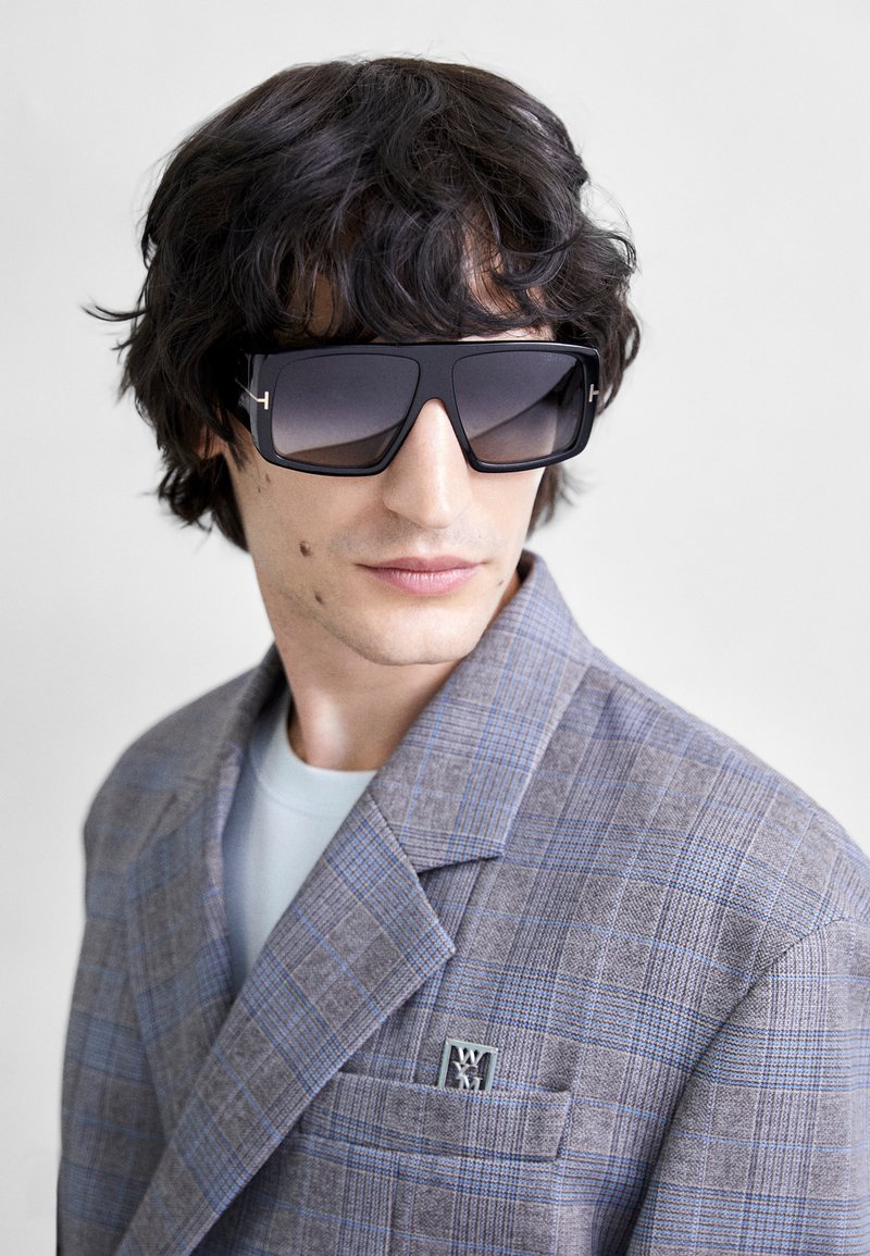 Sorte oversized solbriller med gradientlinser. Grå ternet blazer med subtile blå striber, der har et lommeemblem. Lys blå undertrøje synlig.
