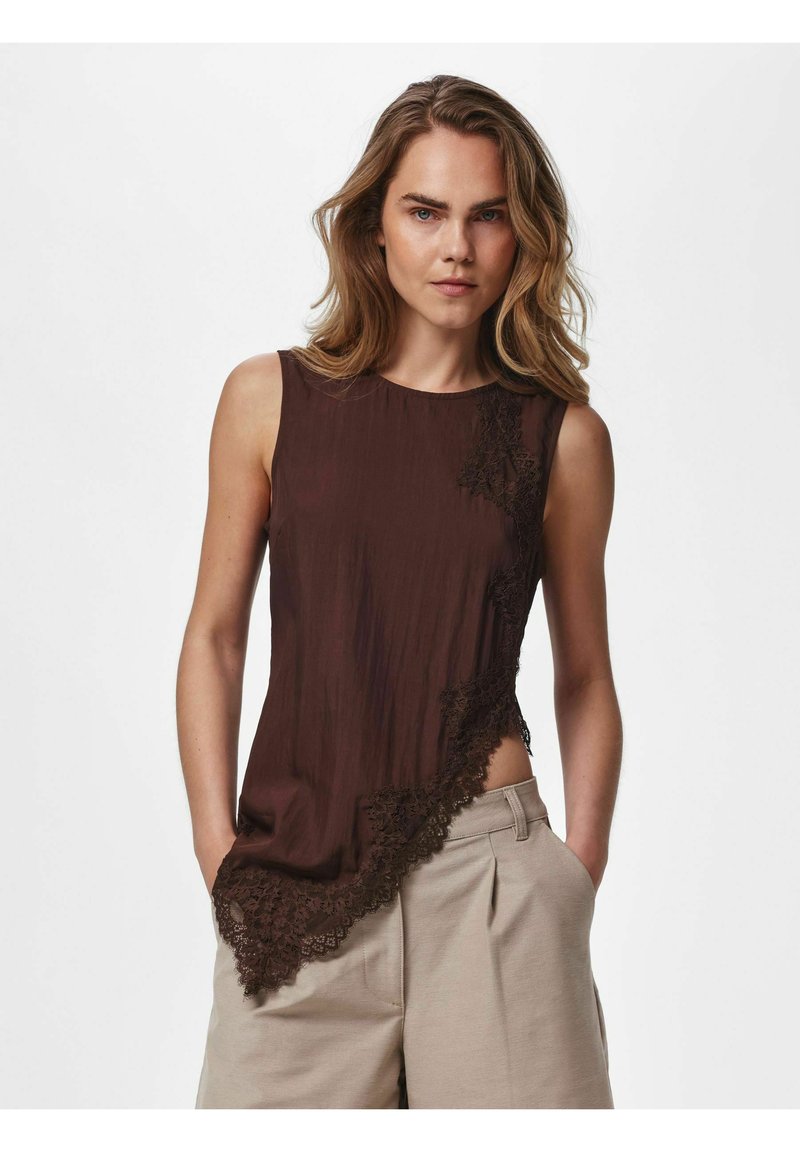 Femme portant un haut marron foncé sans manches avec une bordure en dentelle asymétrique et un pantalon beige taille haute, debout, les mains dans les poches, devant un fond uni.