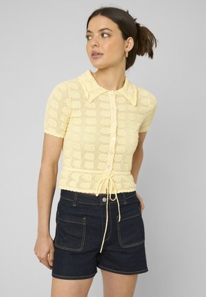 Femme aux cheveux foncés attachés, portant un top en maille jaune pâle à manches courtes boutonné et un short en denim foncé avec poches avant.