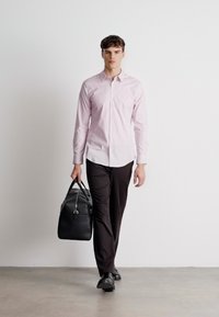 Lichtroze langemouw shirt, zwarte broek, leren zwarte sporttas met riem, en zwarte schoenen; staande op een neutrale achtergrond.