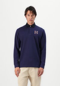 Polo de mangas longas em azul-marinho com gola preta, apresentando um logótipo bordado "H" no peito. Feito de material de algodão suave.