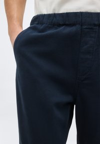 Άνδρας που φοράει σκούρο μπλε παντελόνι με ελαστική μέση, με το αριστερό χέρι στην τσέπη, συνδυασμένο με ένα πουκάμισο λευκό, μισοβαλμένο μέσα.