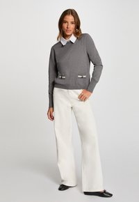 Grijze trui met een V-hals, lange mouwen en twee voorzakken. Combinatie met witte flarde broek en zwarte platte schoenen. Soepele textuur.