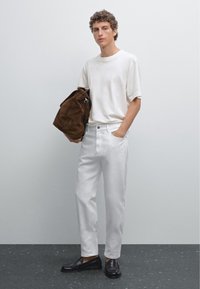 T-shirt bianco in cotone, pantaloni in denim bianchi, borsa a spalla in suede marrone e mocassini neri. Capelli ricci, in piedi contro uno sfondo neutro.