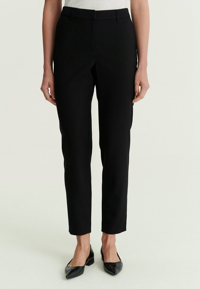 Femme portant un pantalon noir slim et des chaussures noires à talons bas pointues, debout devant un fond clair uni.