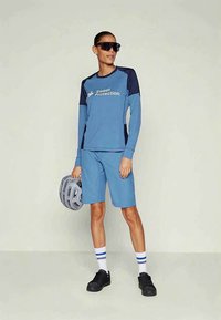 Sieviete zilā garo piedurkņu velosipēdu džersijā un šortos, turot pelēko ķiveri, valkājot melnas sporta kurpes un lielas tumšas saulesbrilles uz neitrāla fona.