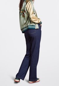Giacca bomber in raso verde con ricamo floreale, abbinata a jeans dritti blu scuro e sneakers verdi con dettagli rossi.