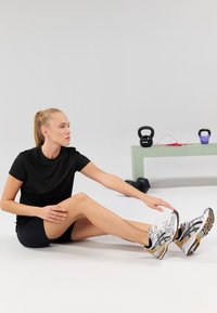 Black athletic t-shirt and shorts, sitting on a white surface. Wearing white and yellow running shoes. Kettlebells and a bench in background.