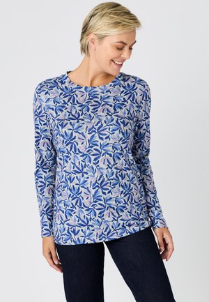 Frau mit kurzen blonden Haaren, die ein langärmliges Oberteil mit blau-lila Blattmustern trägt, lächelt und sieht nach unten.