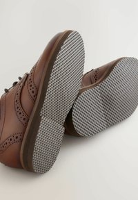 Chaussures en cuir marron avec des détails en brogue décoratifs et des semelles en caoutchouc grises présentant un motif texturé. Lacets et design de bout rond visibles.