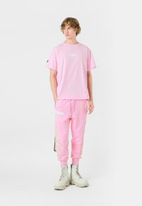 Camiseta rosa con un parche rectangular blanco, combinada con joggers rosas a juego que cuentan con acentos laterales brillantes y un bolsillo con cremallera. Botas blancas de suela gruesa.