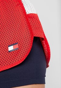 Rotes Mesh-Sporttrikot mit weißen Akzenten und einem kleinen Logo-Patch am Saum. Über einer marineblauen Basislage getragen, zeigt es einen atmungsaktiven Stoff.