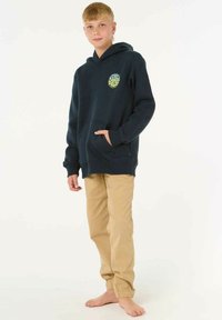 Sudadera con capucha de color azul marino con bolsillo delantero y un logo circular amarillo, combinada con pantalones de color beige con puños elásticos, modelo de pie descalzo.