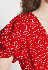 Vestido floral rojo con mangas cortas abullonadas. Presenta un escote en V y pequeñas flores blancas sobre un fondo vibrante, hecho de una tela ligera.