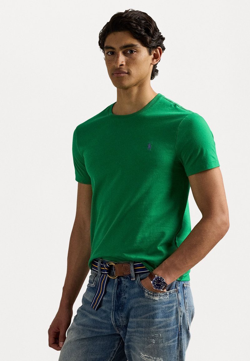 T-shirt de algodão verde com decote redondo e mangas curtas, apresentando um pequeno logotipo roxo. Usado com jeans claros desgastados e um cinto listado.