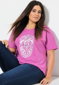 Różowa koszulka z krótkim rękawem z szczegółowym graficznym wizerunkiem srebrnego lwa z przodu, zestawiona z ciemnoniebieskimi dżinsami.