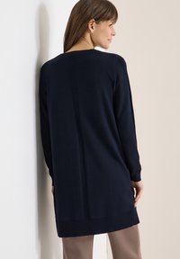 Femme portant un long pull bleu marine avec une bande côtelée verticale dans le dos, associée à un pantalon marron clair, face à un mur blanc.