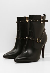 Bottes en cuir noir à chevilles avec des bouts pointus, des accents en clous dorés, deux sangles décoratives et une fermeture à glissière sur le côté. Talons aiguille.