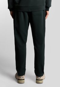 Donkergroene trainingsbroek met een zachte textuur en elastische tailleband, met een detail van een achterzak. G gedragen met beige sportschoenen.