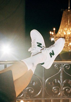 Person lounging with legs raised, wearing white New Balance sneakers and white socks, ornate iron railing and chandelier in the background at night.