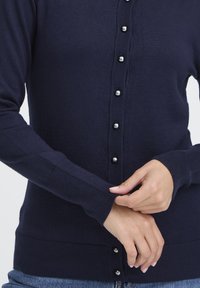 Marineblauwe cardigan gemaakt van een zachte brei stof, met een knopenfront met metalen accenten en geribbelde manchetten en zoom.
