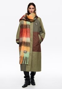Oliwkowy płaszcz z brązowymi akcentami i logo. Oversizowy wielokolorowy szalik w kratę z frędzlami. Czarne dopasowane spodnie i kozaki sięgające kolan.
