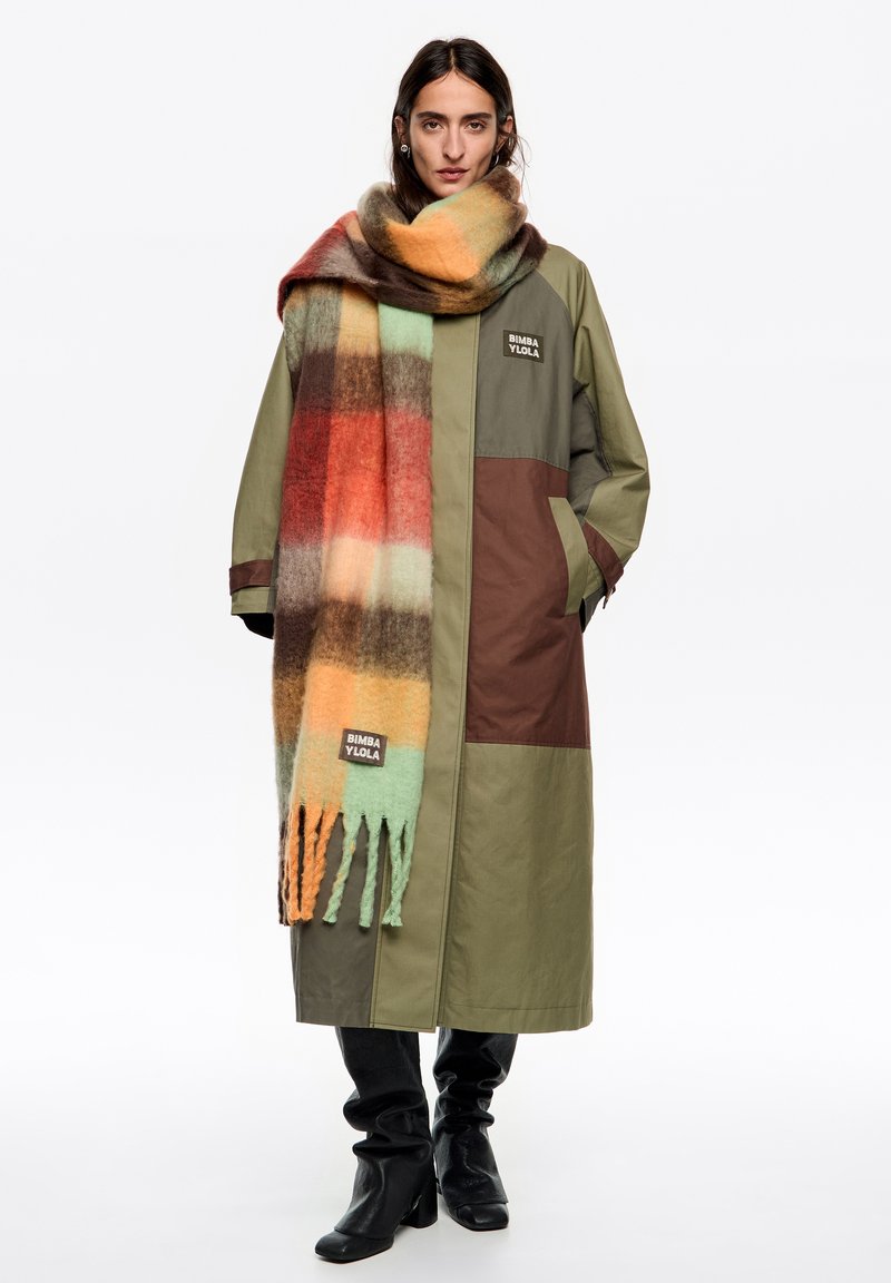 Oliwkowy płaszcz z brązowymi akcentami i logo. Oversizowy wielokolorowy szalik w kratę z frędzlami. Czarne dopasowane spodnie i kozaki sięgające kolan.