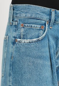 Detailný záber svetlomodrých džínsových nohavíc s vreckom, medenými nitmi a viditeľným šitím na čiernom pozadí.