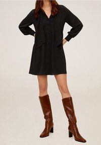 Robe noire à manches longues avec un devant boutonné et une ceinture à la taille. Associée à des bottes en cuir marron à talons montant jusqu'aux genoux. Texture lisse.