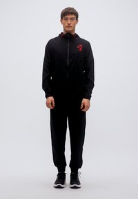 Μαύρη αθλητική φόρμα με μπουφάν με φερμουάρ και ελαστικό παντελόνι. Διαθέτει κόκκινες λεπτομέρειες και λογότυπο στο στήθος. Ομαλό ύφασμα, εφαρμοστή σχεδίαση.