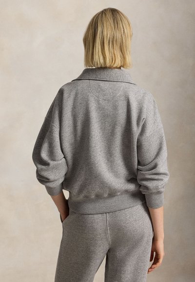 Persoon met kort blond haar, gekleed in een grijze trui met lange mouwen en bijpassende grijze broek, gezien van achteren tegen een neutrale achtergrond.
