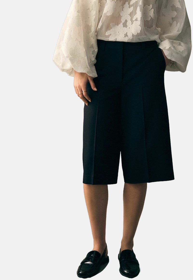 Svarta skräddarsydda shorts med veck, kombinerat med en genomskinlig vit blus med blommönster och svarta loafers. Tyget verkar glatt och strukturerat.