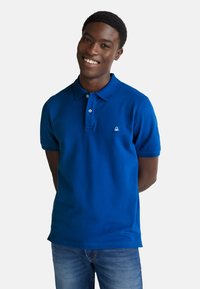 Usmívající se mladý muž s krátkými vlasy v modrém polo tričku a džínách, stojící s rukama za zády na bílém pozadí.