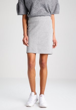 Femme portant une jupe patchwork gris clair, un pull gris à manches courtes et des chaussures de sport blanches, debout sur un fond uni.