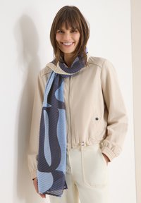 Femme souriante portant une veste beige et un pantalon crème avec une écharpe à motifs bleu et marine, debout contre un mur clair.