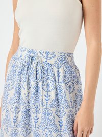 Bantul cu broderie florală albastră pe o fustă dintr-un material ușor. Prezintă detalii pliate pentru formă. Fundalul este simplu, alb.