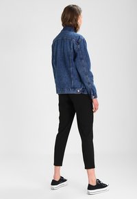Persona de pie, de espaldas, vistiendo una chaqueta de mezclilla azul, pantalones negros cortos y zapatillas de lona negras sobre un fondo liso.