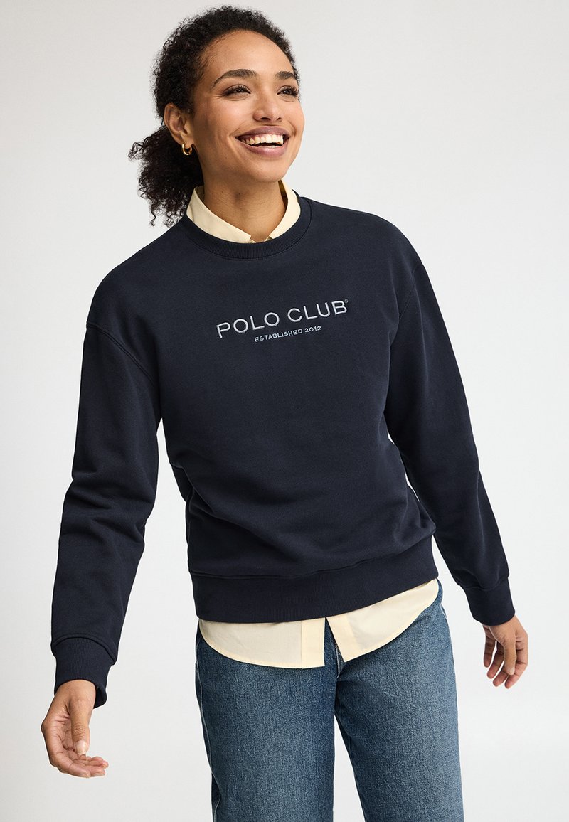 Smilende kvinde med krøllet hår iført en navy "Polo Club" sweatshirt over en lysegul skjorte og blå jeans, stående mod en ensfarvet hvid baggrund.