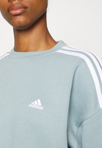 Felpa azzurra chiara con strisce bianche sulle spalle e un piccolo logo Adidas bianco sul petto. Realizzata in tessuto morbido e testurizzato.