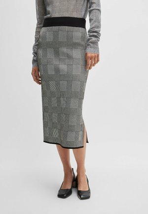 Femme portant une jupe midi à carreaux gris avec une taille noire, un haut à manches longues gris assorti et des chaussures à petits talons noires, se tenant sur un fond blanc.