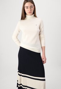 Pull à col roulé crème en tricot doux, associé à une jupe plissée marine avec des rayures crème et noires. Marquage minimal sur le pull.