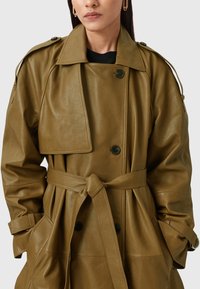 Olijfgroen leren trenchcoat met een riem in de taille, brede kraag, oversized mouwen en twee voorkruiszakken, voorzien van zwarte knoopsluitingen.
