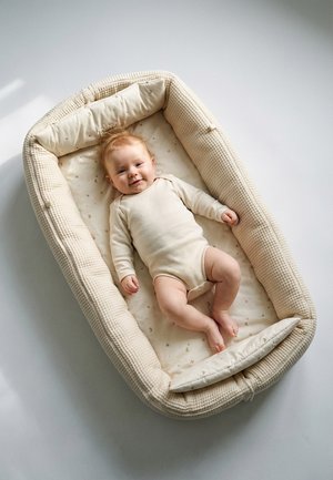 Lächelndes Baby in einem cremefarbenen Strampler, liegend in einem weichen, gepolsterten beigen Baby-Nest mit Mond- und Sternenmuster auf der Matratze.