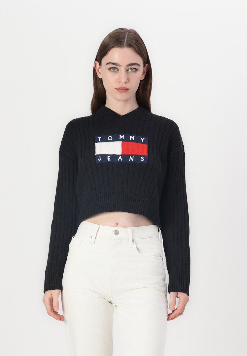 Černý žebrovaný krátký svetr s obdélníkovým vyšívaným logem "TOMMY JEANS" v bílé, červené a modré barvě. Nosit s bílými džíny.