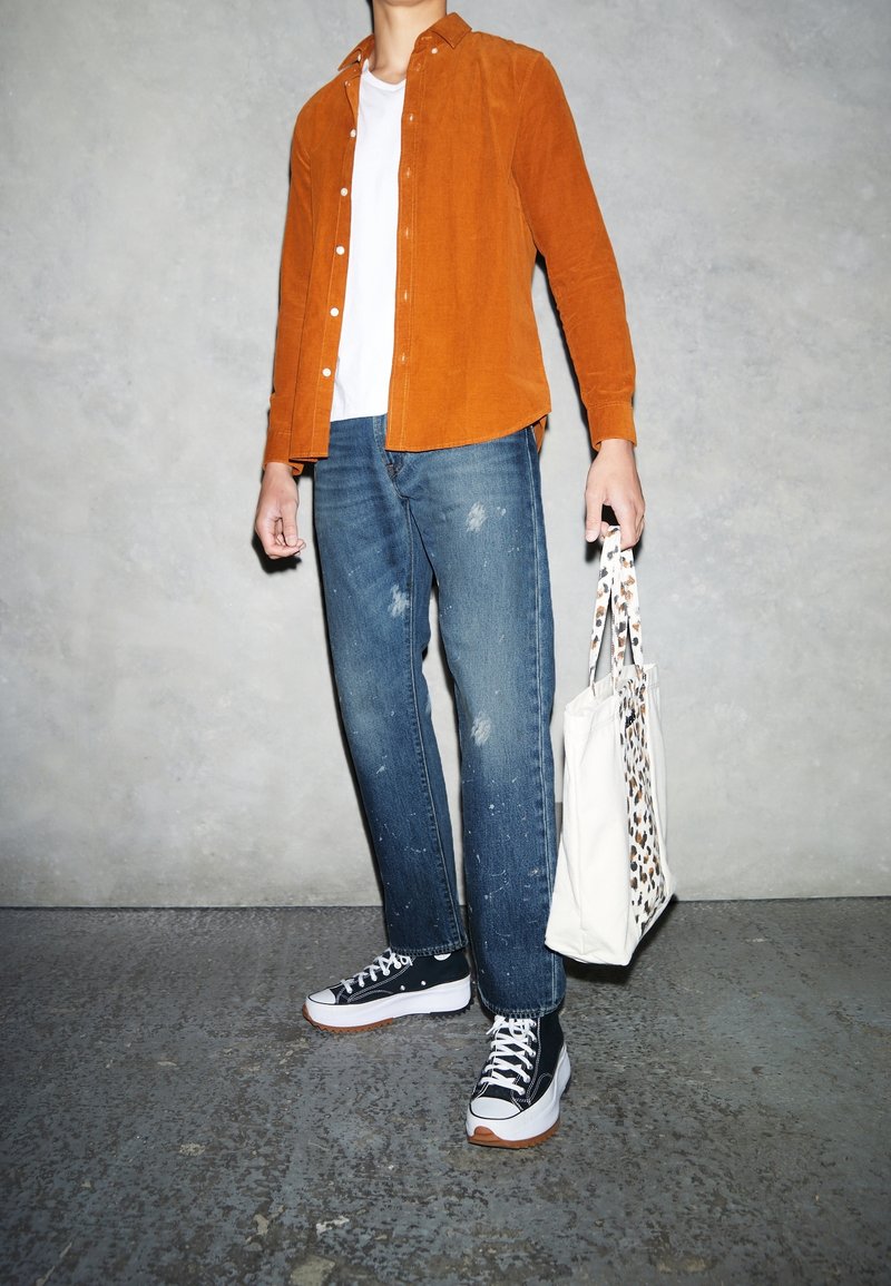 Personne portant une chemise orange à boutons, un t-shirt blanc, un jean bleu déchiré, des baskets noires et blanches, tenant un sac fourre-tout blanc avec des poignées imprimées léopard.