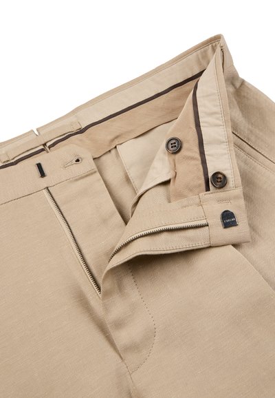 Pantalon beige avec une fermeture éclair partiellement ouverte et une double fermeture par boutons, dévoilant la ceinture intérieure avec un détail de bordure foncée.