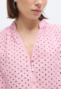 Naine, kellel on rõngakujulised kõrvarõngad ja heleroosa silmkoelise pluus, millel on väike volditud krae ja kolm nööpi kaelusel.